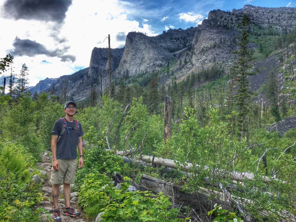 Tim standing on a trail in a forested area with cliffs in the background. 